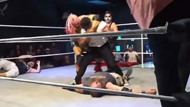 Wrestling match inside a ring; a masked wrestler with white face paint stands over a fallen opponent while another wrestler kneels nearby and a fourth person lies on the mat outside the ring.