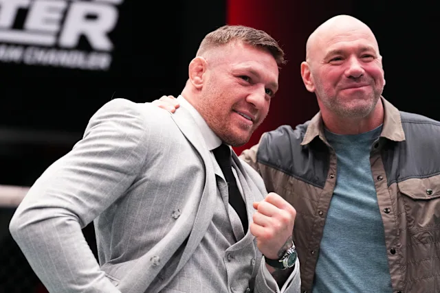 Two men posing for a photo together; one in a light gray suit and tie, the other in a brown jacket and blue shirt, both smiling at the camera.