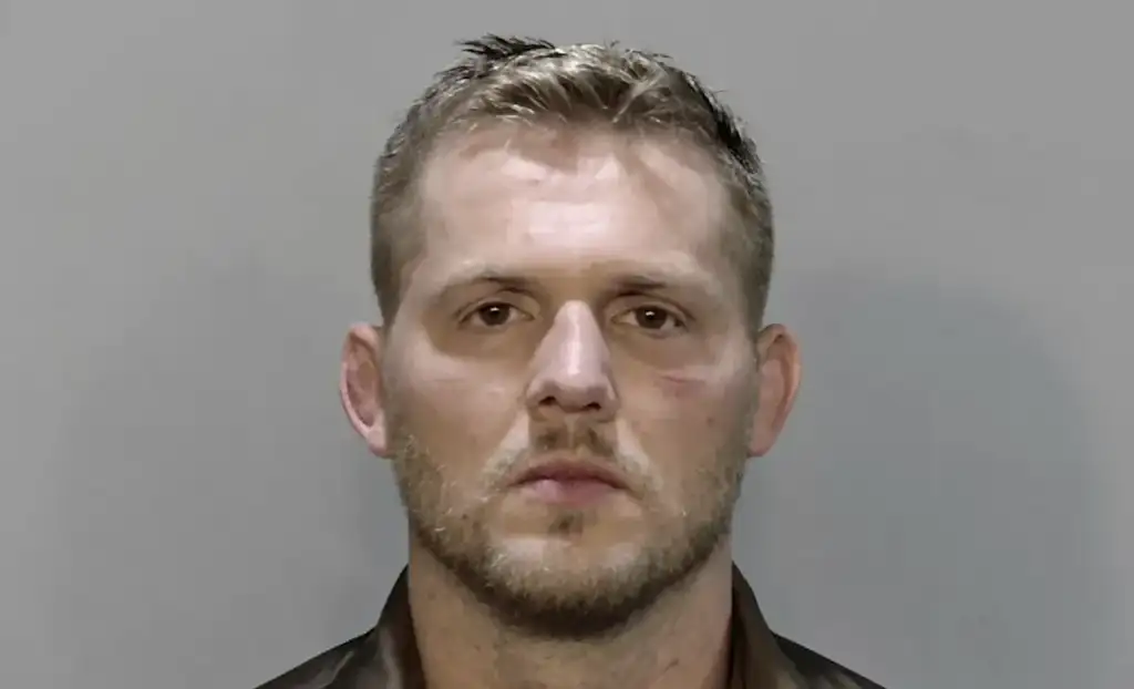 Mugshot of a man with short light brown hair and stubble, facing forward against a gray backdrop.