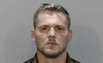 Mugshot of a man with short light brown hair and stubble, facing forward against a gray backdrop.