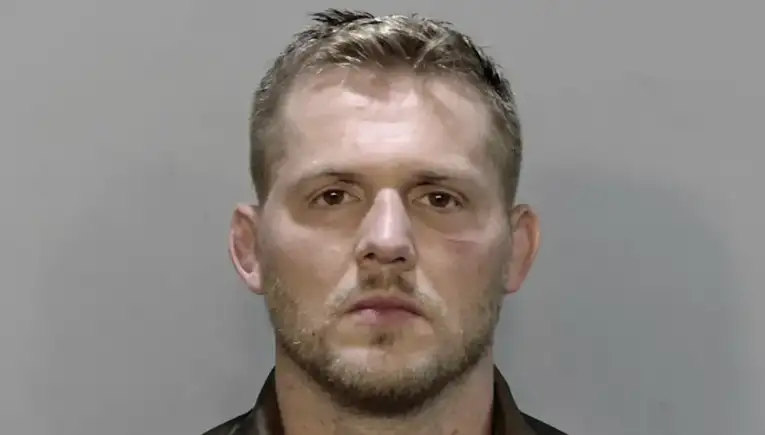 Mugshot of a man with short light brown hair and stubble, facing forward against a gray backdrop.