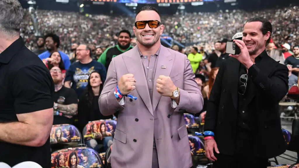 Man in a lavender suit and orange sunglasses poses with fists up at a crowded stadium event, American flag pin on lapel.