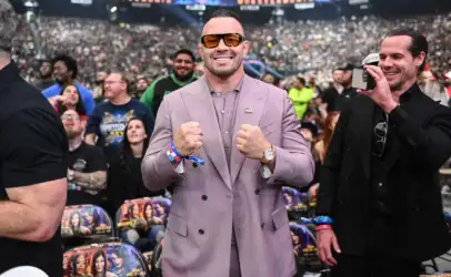 Man in a lavender suit and orange sunglasses poses with fists up at a crowded stadium event, American flag pin on lapel.