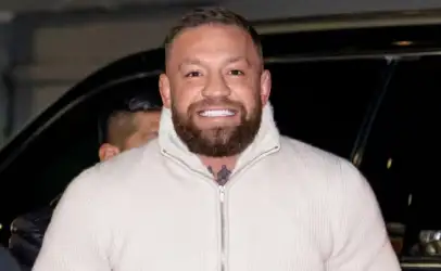 Smiling man with a trimmed beard wearing a cream zip-up sweater outdoors at night in front of a dark car of background.