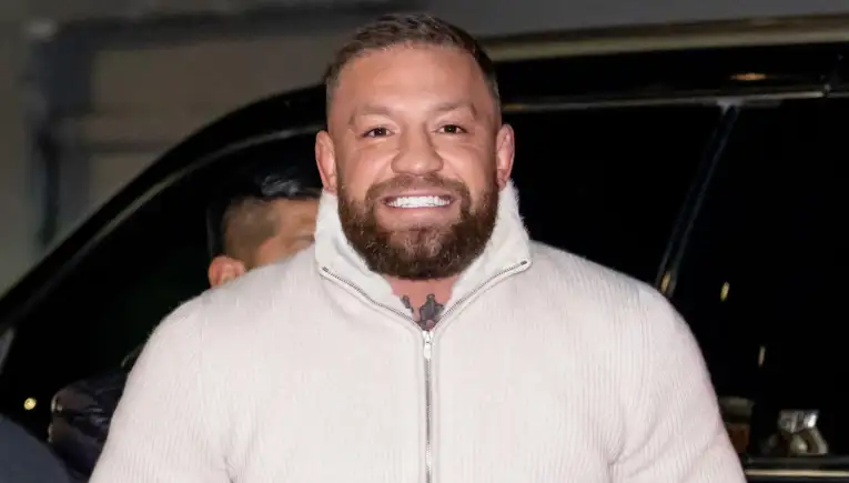 Smiling man with a trimmed beard wearing a cream zip-up sweater outdoors at night in front of a dark car of background.
