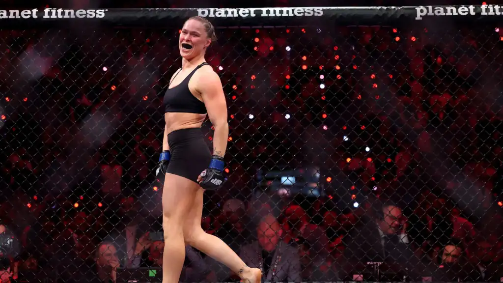 crop-39132055_121bd2 - AfroCombat Female MMA fighter in a black sports bra and shorts with blue gloves, celebrating inside a fenced cage with a cheering crowd in the background.