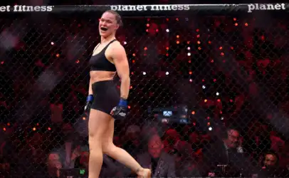 Female MMA fighter in a black sports bra and shorts with blue gloves, celebrating inside a fenced cage with a cheering crowd in the background.