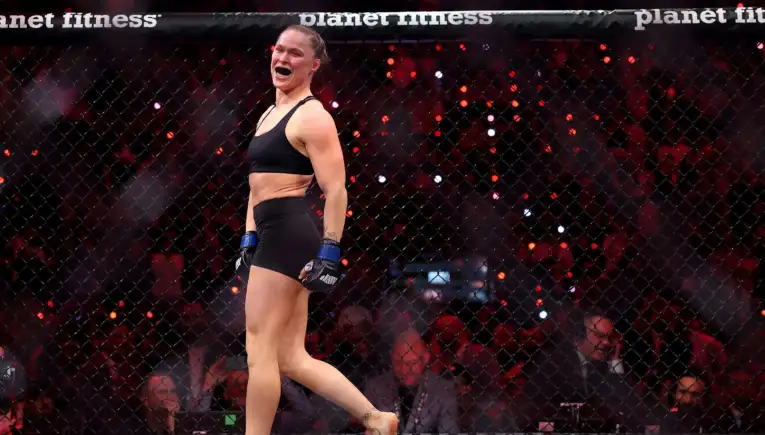 Female MMA fighter in a black sports bra and shorts with blue gloves, celebrating inside a fenced cage with a cheering crowd in the background.