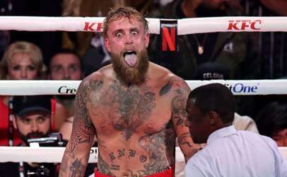 Tattooed, bearded boxer with mouth open shouting in the ring as a referee and crowd watch nearby.