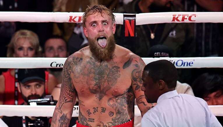 Tattooed, bearded boxer with mouth open shouting in the ring as a referee and crowd watch nearby.