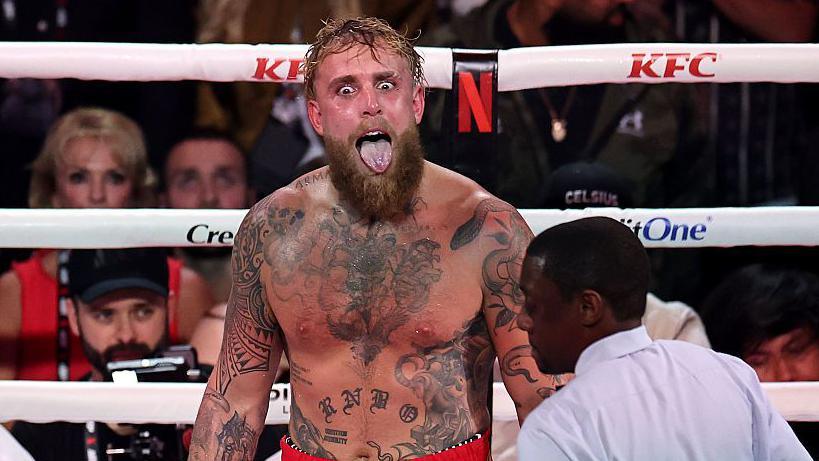 Tattooed, bearded boxer with mouth open shouting in the ring as a referee and crowd watch nearby.