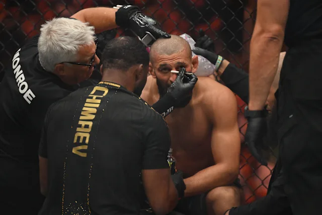 e1e9f76b00a6fd1cc4081f5056c8cfe8 - AfroCombat MMA fighter sits shirtless in the cage as his cutman and teammates tend to a facial injury near his eye.