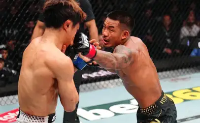 Two shirtless mixed martial artists spar inside a cage; the fighter on the right throws a punch toward the opponent's head as the other braces with gloves up.