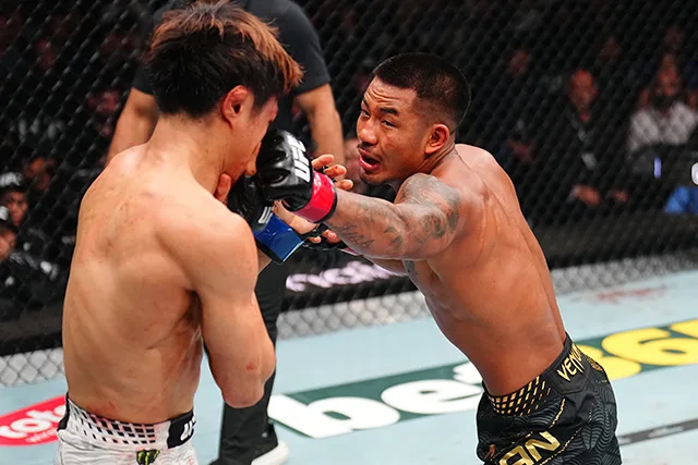 Two shirtless mixed martial artists spar inside a cage; the fighter on the right throws a punch toward the opponent's head as the other braces with gloves up.