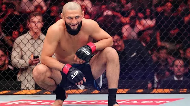 UFC fighter crouches in the octagon, smiling, wearing red gloves and black shorts with the crowd behind the cage.