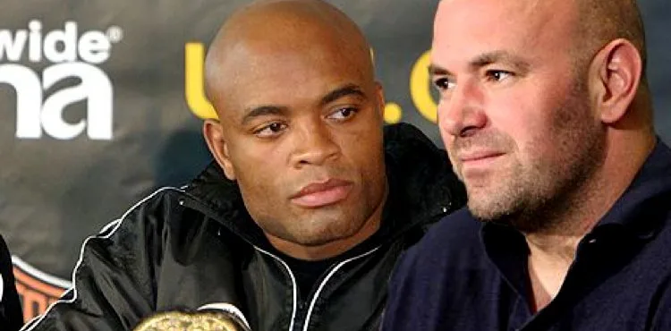 Two men at a press conference, one bald in a black jacket and the other with short hair and a stubbled chin; a boxing belt is visible in the foreground.
