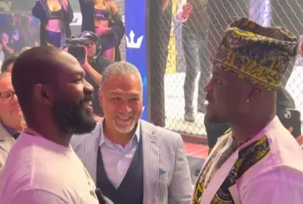 f706ce91084fbb1e653b17977a057413 - AfroCombat Three men stand and chat inside a fenced sports arena, smiling at each other.
