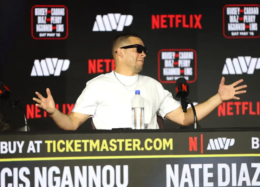 Man wearing a white shirt and sunglasses speaks at a press conference with Netflix logos in the background, gesturing with hands.