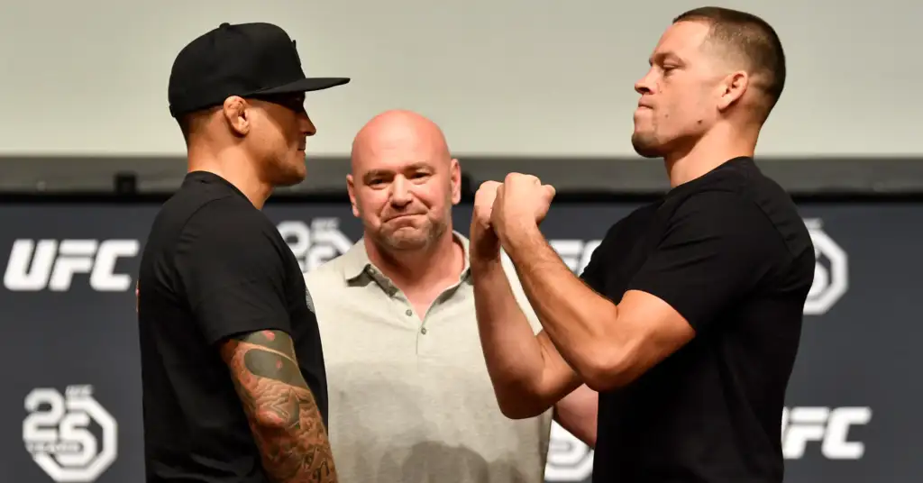 Two men in black shirts face off in a weigh-in, fists raised toward each other, with a smiling official between them against a UFC backdrop.