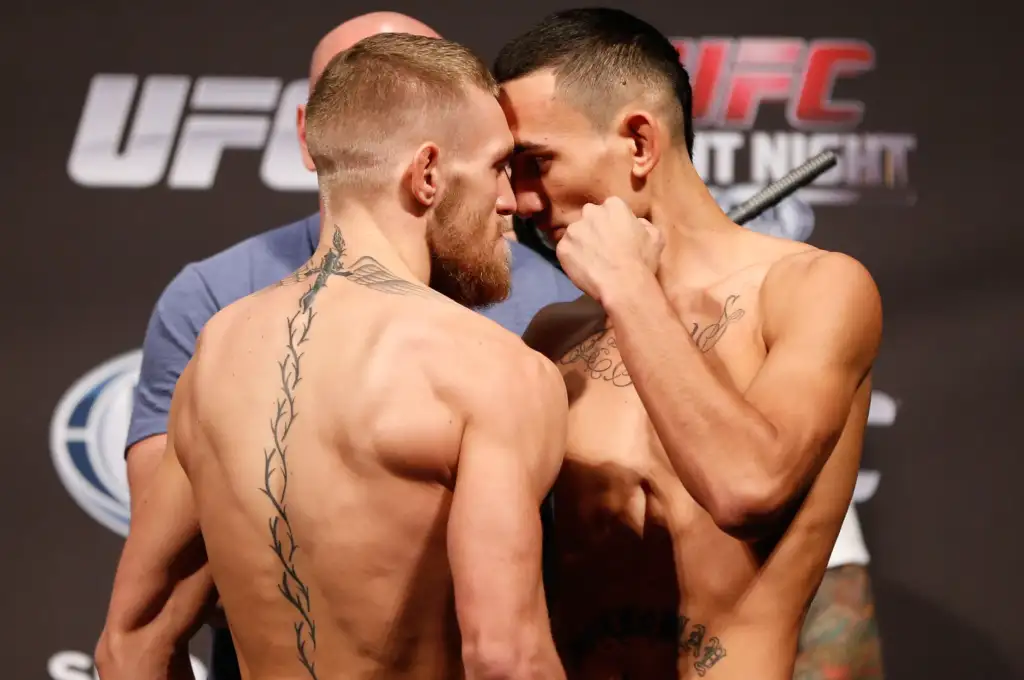 Two shirtless men face off in a UFC weigh-in, foreheads touching, with tattoos visible on their backs and arms and a judge or staffer in the background.