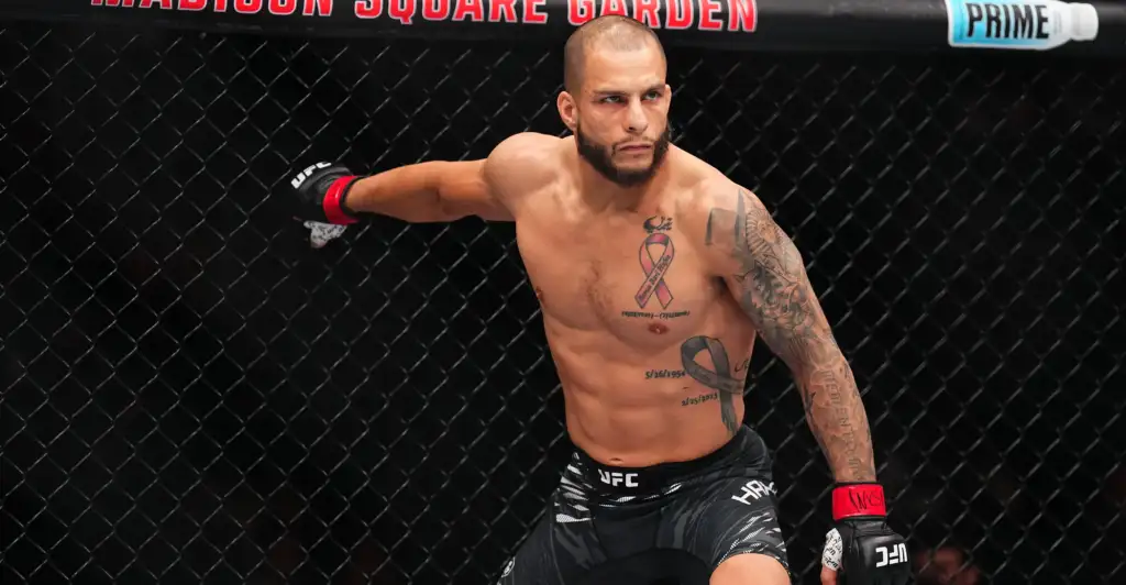 Male MMA fighter inside the UFC octagon, shirtless, extending his arm against the chain-link fence, wearing black shorts with UFC branding and red Wristwraps, tattoos visible on his chest and arm.