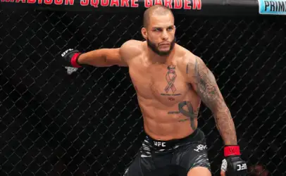 Male MMA fighter inside the UFC octagon, shirtless, extending his arm against the chain-link fence, wearing black shorts with UFC branding and red Wristwraps, tattoos visible on his chest and arm.