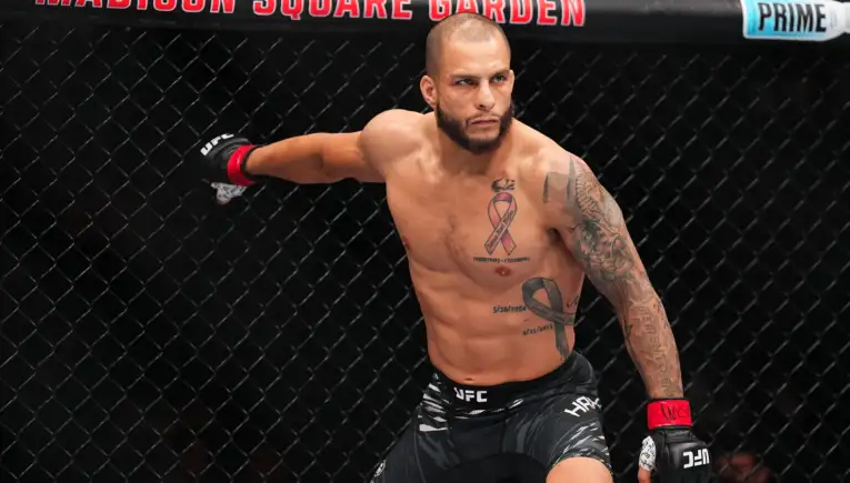 Male MMA fighter inside the UFC octagon, shirtless, extending his arm against the chain-link fence, wearing black shorts with UFC branding and red Wristwraps, tattoos visible on his chest and arm.