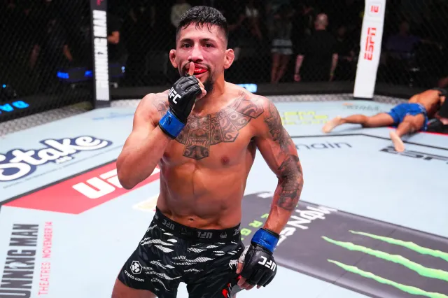 Male mixed martial artist in the octagon, shirtless and gloved, touching his lips with his glove in a victory pose after a fight.