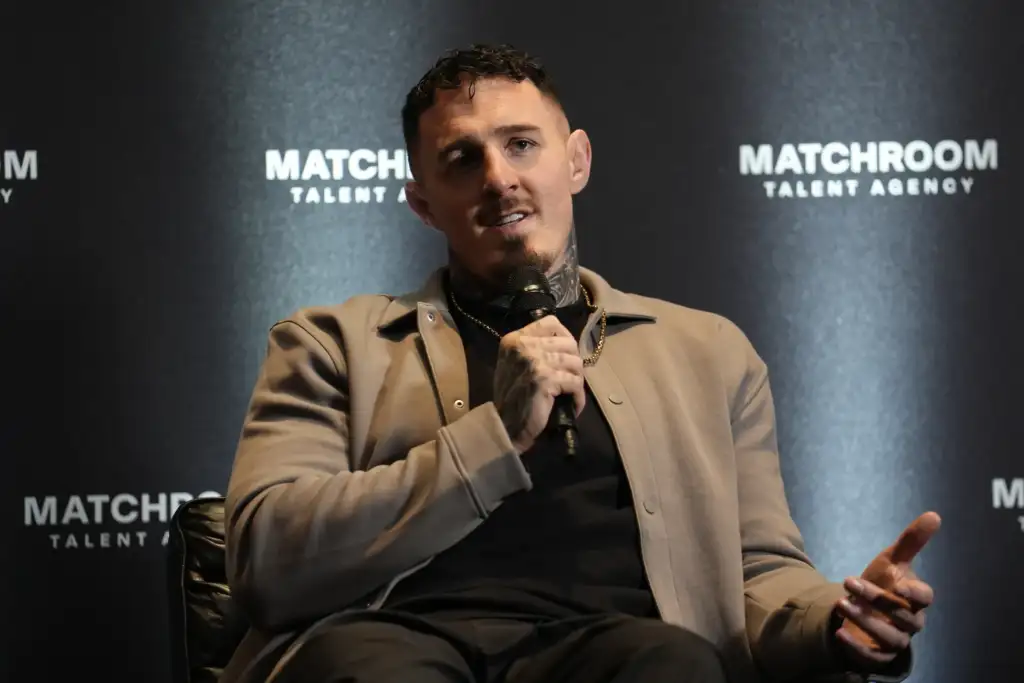 Man speaking into a handheld microphone on a stage with a dark backdrop that says 'Matchroom Talent Agency' behind him, wearing a beige jacket and black shirt, with visible neck tattoo.
