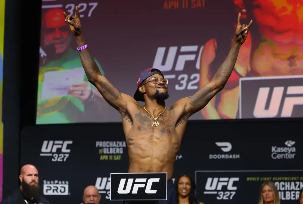 UFC fighter with arms raised on stage at a weigh-in, logos and 'UFC 327' visible on the backdrop behind him