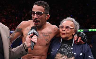 UFC fighter with tattoos and glasses answers questions into a microphone, standing with an elderly woman in a navy jacket inside the arena.