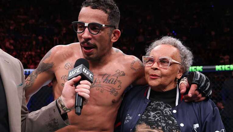 UFC fighter with tattoos and glasses answers questions into a microphone, standing with an elderly woman in a navy jacket inside the arena.