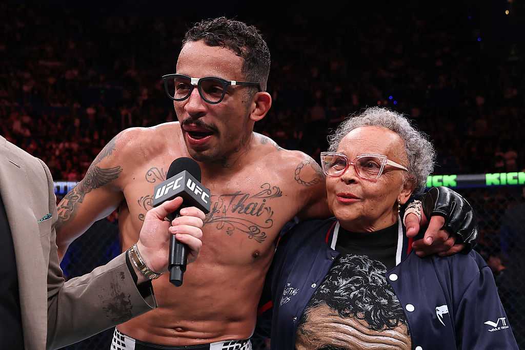 UFC fighter with tattoos and glasses answers questions into a microphone, standing with an elderly woman in a navy jacket inside the arena.