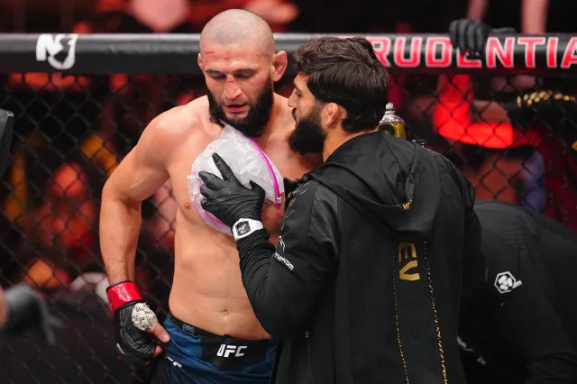 Two mixed martial artists inside the cage after a match: one shirtless with a shaved head and red gloves, the other in a black jacket leaning in close beside him.
