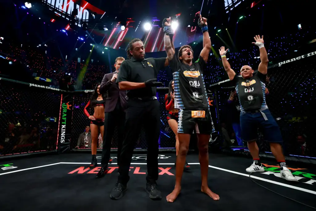 Moment of victory in a mixed martial arts match: fighters raise arms in the cage as the referee stands nearby, crowd lights shining overhead.