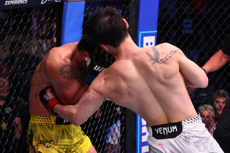 Two male UFC fighters exchange punches inside the octagon, one wearing yellow shorts and the other in white trunks.