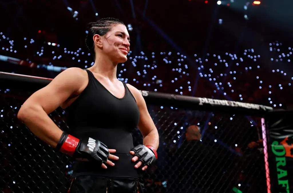 Female mixed martial artist stands in the cage, wearing a black outfit and red gloves, looking toward the crowd with a smile.