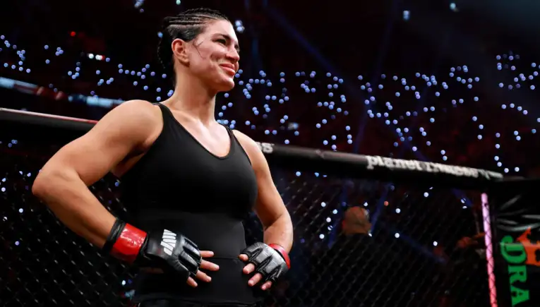 Female mixed martial artist stands in the cage, wearing a black outfit and red gloves, looking toward the crowd with a smile.