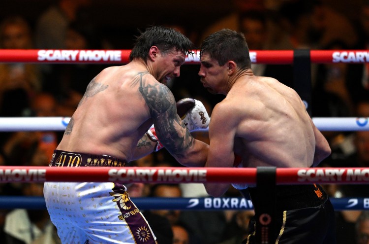 Two shirtless male boxers exchange punches in a ring during a match, ropes labeled 'SCARY MOVIE' and a blurred crowd in the background.