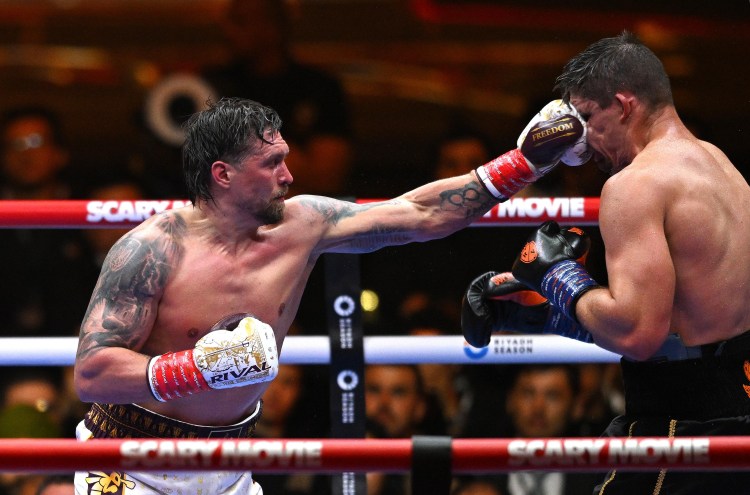 Two shirtless boxers in a ring exchanging a punch; the left fighter lands a hook to the opponent's face as the crowd watches.Recognizable ropes and banners frame the action, capturing a tense moment in the bout.
