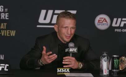 MMA fighter in a black suit speaks into a microphone at a UFC press conference, with UFC banners behind and a championship belt on the table nearby.