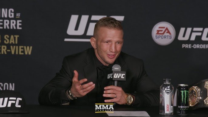 hq720 19 - AfroCombat MMA fighter in a black suit speaks into a microphone at a UFC press conference, with UFC banners behind and a championship belt on the table nearby.