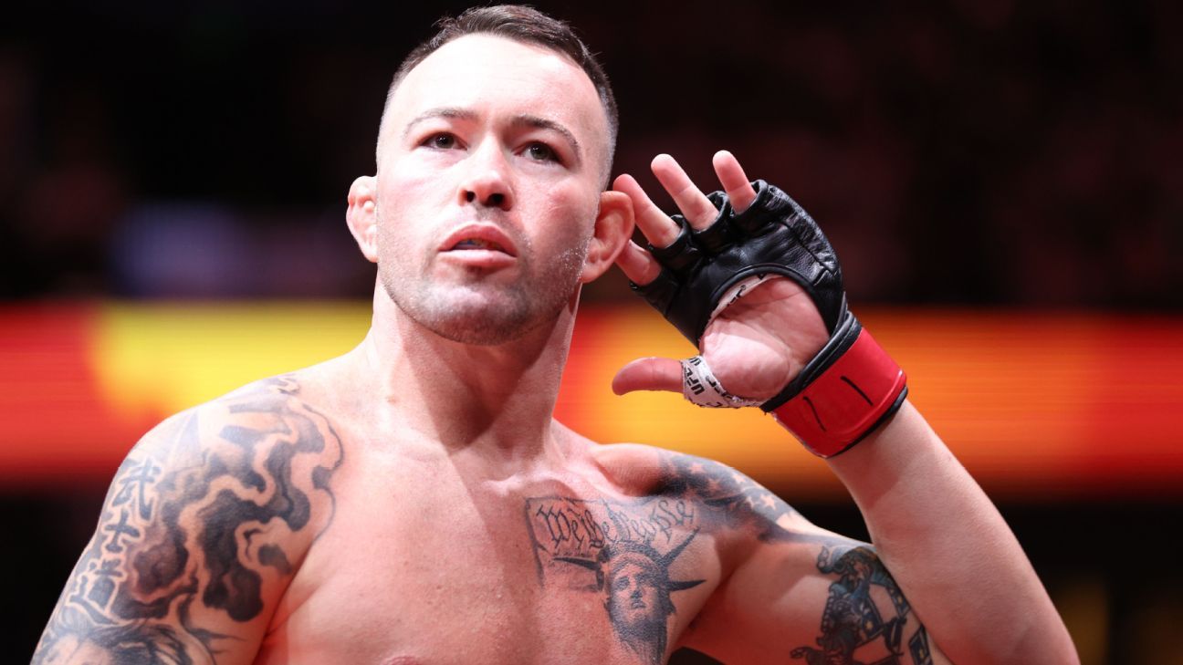 Male mixed martial artist in a black glove and red wristband raises his right hand to his ear, shirtless with tattoos on his chest and arms.