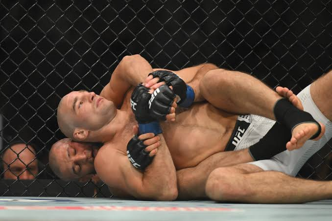 Two mixed martial artists grapple on the mat inside a chain-link cage, one pinning the other with a hold and wearing UFC gloves.