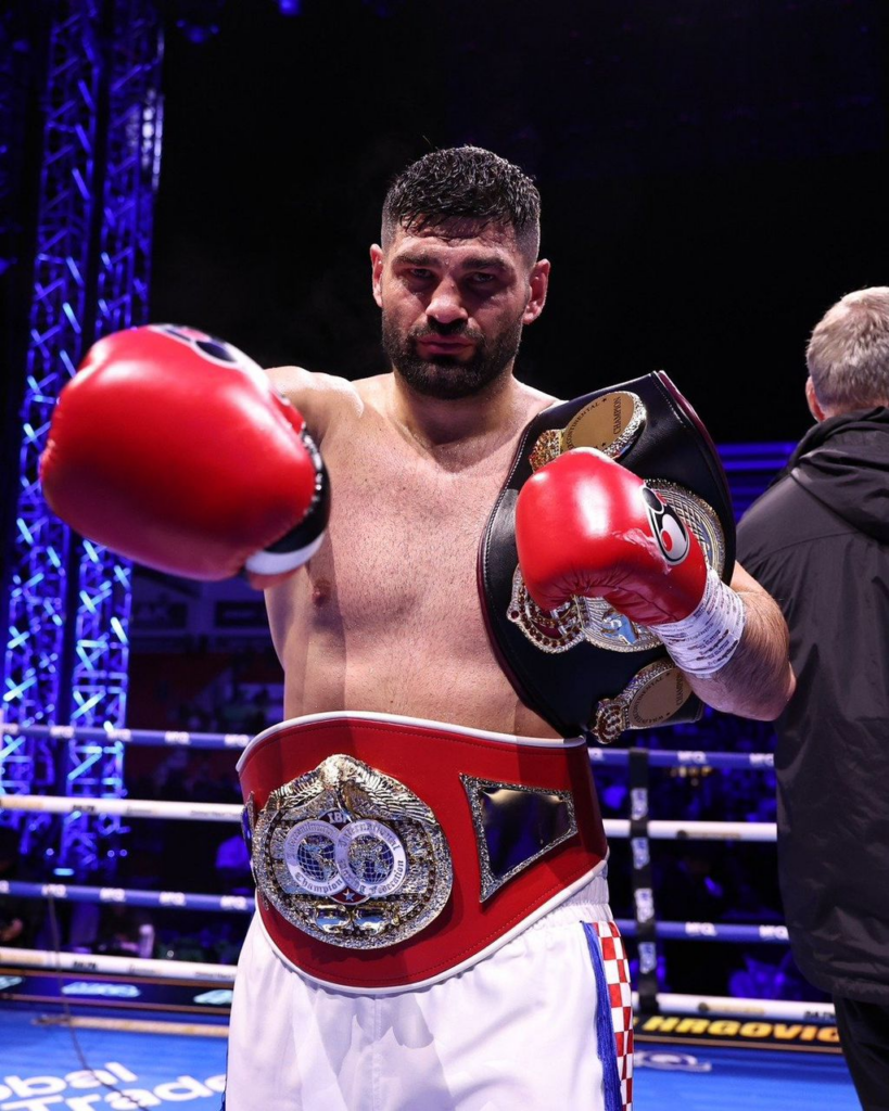 Boxer in the ring wearing multiple championship belts, arms raised with red gloves.