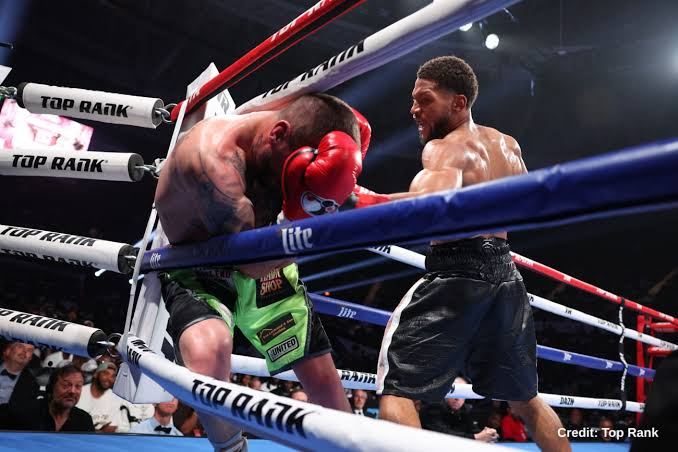 Two boxers exchange punches in a ring, one wearing red gloves and green shorts, the other in black shorts, as they trade blows amid blue and white ropes labeled Top Rank.