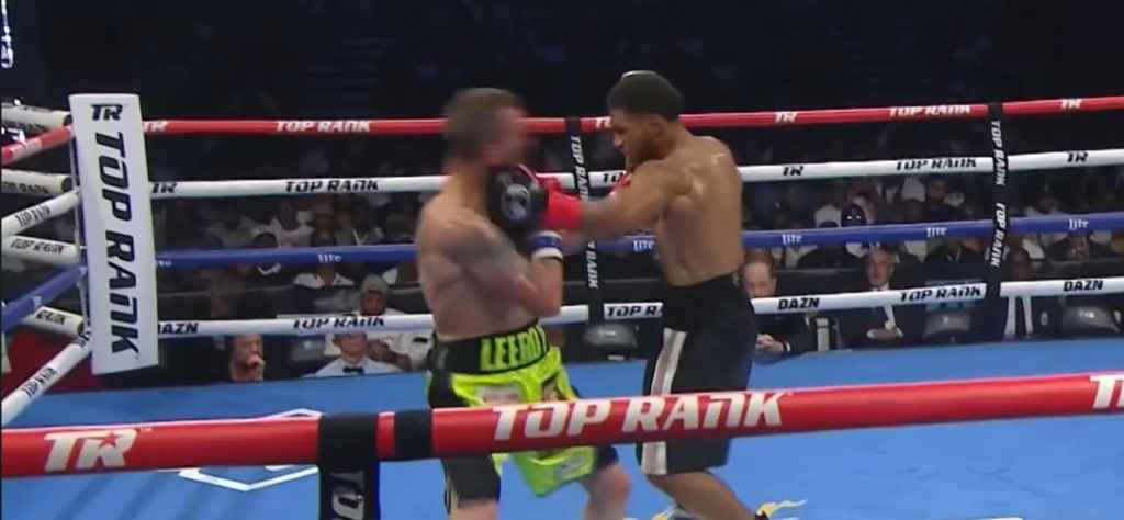 Two shirtless boxers clash in a ring as one lands a punch to the other's head, spectators watching from the stands.