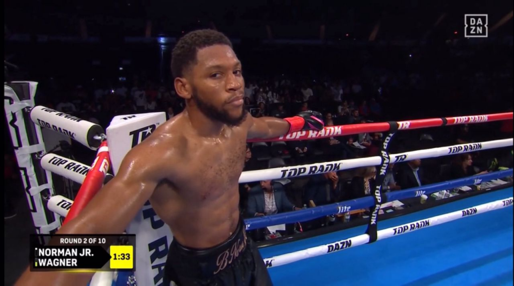 Boxer rests against the ropes in a boxing ring during a DAZN broadcast, scoreboard reads Round 2 of 10 with Norman Jr. Wagner and a 1:33 timer on screen.