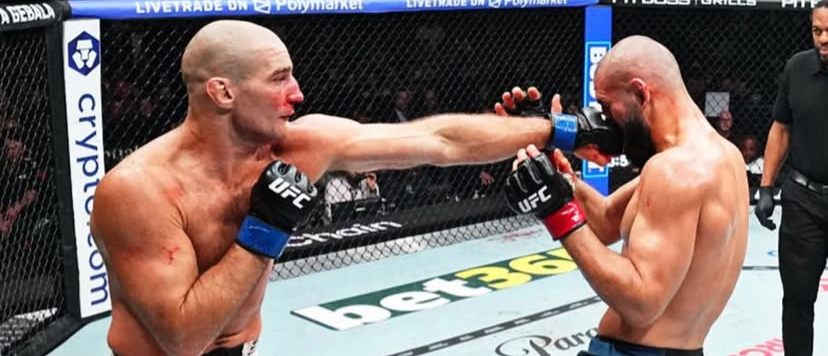 Two UFC fighters in a cage exchange punches; one lands a right hook to the other’s face, blood visible on the nose area.