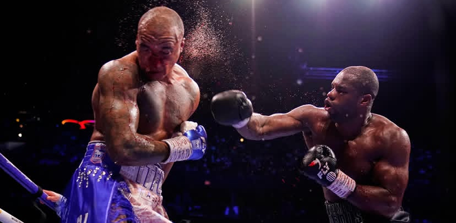 Two muscular boxers exchange punches in a dimly lit boxing ring during a match, with one landing a punch on the other.